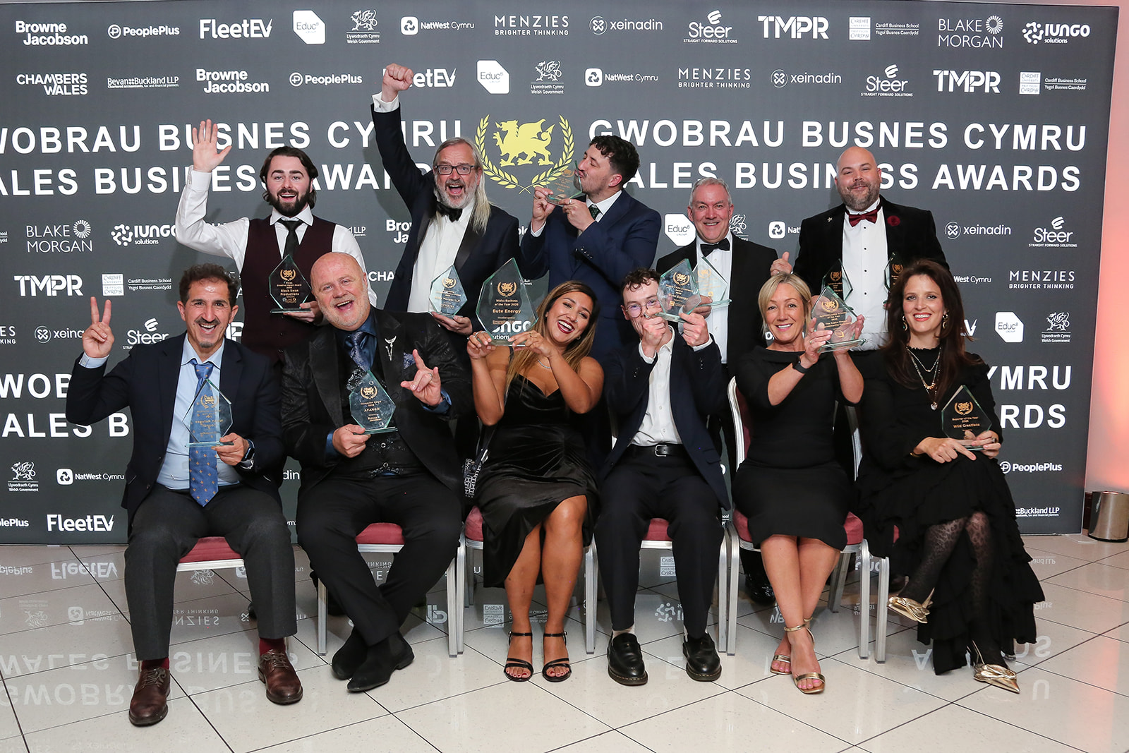 a group of people in formal wear holding up awards in front of a Wales business awards backdrop